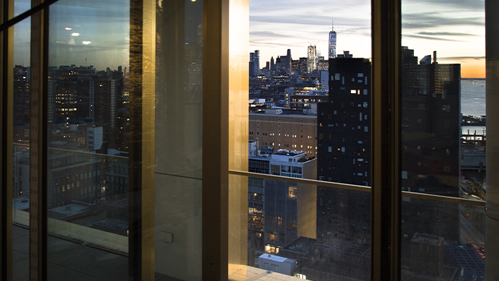 Interno di un grattacielo di New York, con vista dello skyline al tramonto. In primo piano una grande vetrata con sistemi in alluminio dorati ad elevato isolamento acustico.