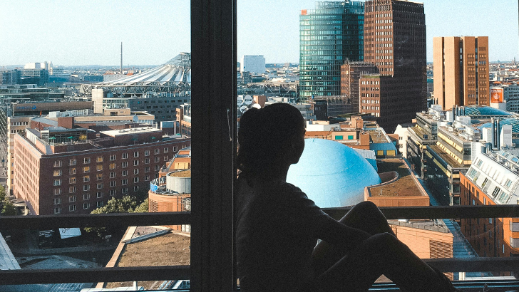 Interno di un stanza di grattacielo, con in primo piano una ragazza con lo sguardo rivolto verso l'esterno, con vista sul paesaggio urbano di una metropoli